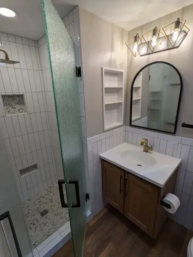 Bathroom with white tiled shower, wood vanity, and black-framed mirror.