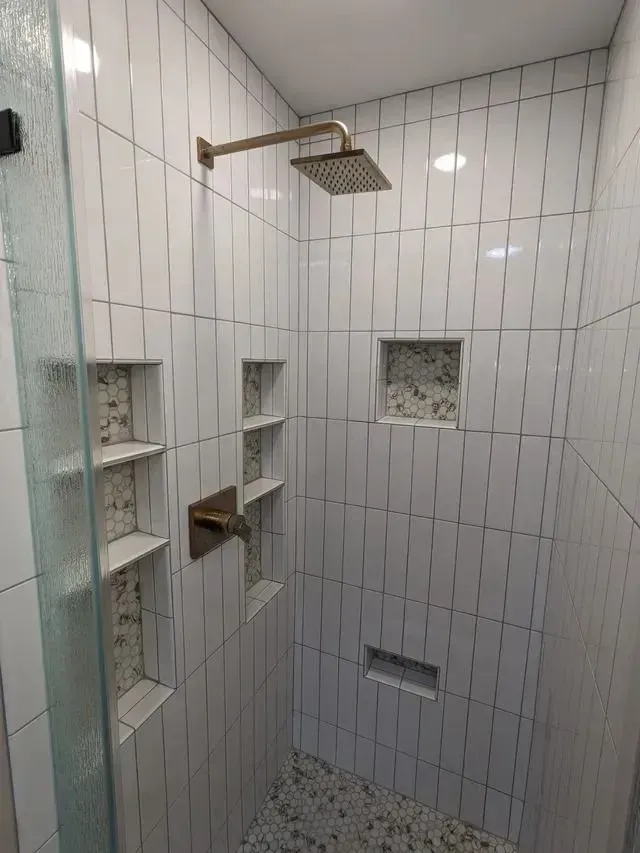 White-tiled shower with multiple built-in shelves. Gold showerhead and fixtures. Pebble floor. Glass shower door.