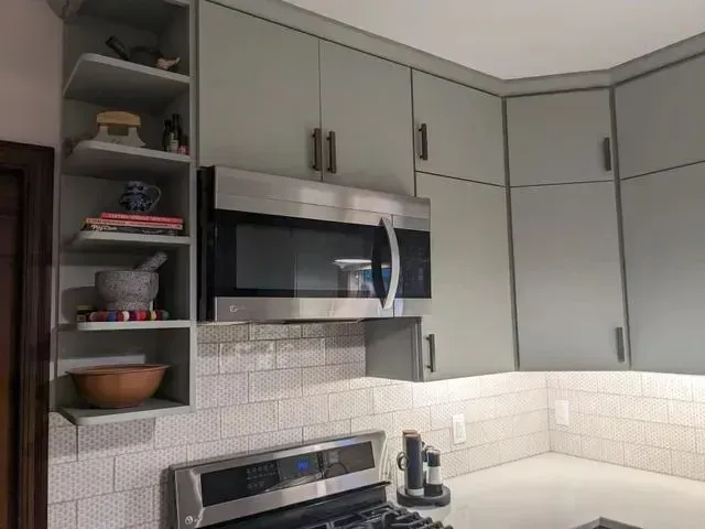 Kitchen cabinets in a sage green color, microwave, and open shelves with kitchen items. White backsplash.