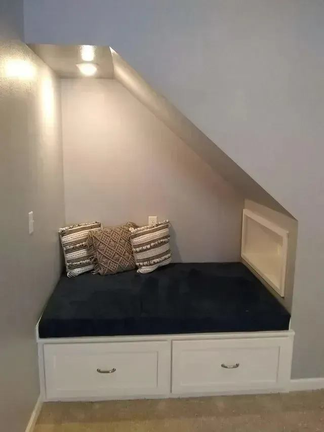 Built-in reading nook under stairs with blue cushion, pillows, white drawers, and recessed lighting.
