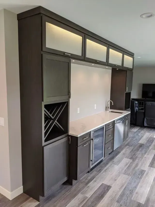 Dark brown built-in bar with upper cabinets, sink, wine rack, mini-fridge, and dishwasher against a gray wall.