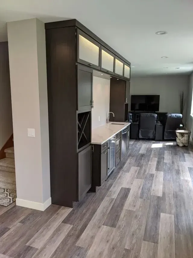 Dark brown custom bar with overhead cabinets, sink, and mini-fridge in a gray-floored living area.