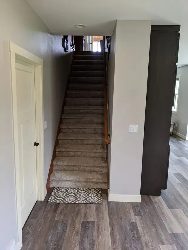 Staircase with carpeted steps, wooden handrail, leading up. A white door and gray walls are present.