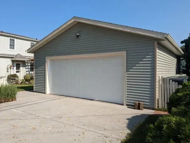Gray-sided garage with white garage door on a concrete driveway, next to a wooden fence, under a blue sky.
