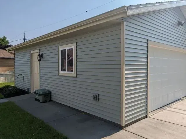 Blue-sided garage with beige trim, a white window, and a closed garage door. Sunny day.
