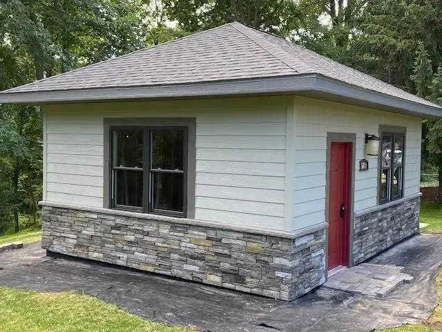 Small square building with stone veneer base, light siding, dark windows, and a red door.