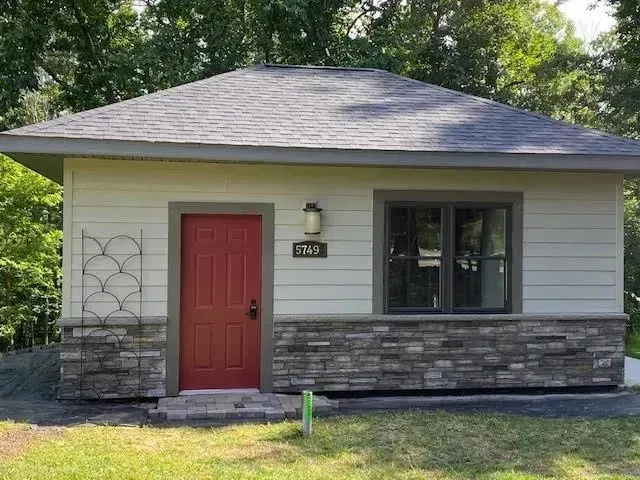 Small beige building with red door, stone facade, and gray roof. Window, outdoor light, and greenery visible.