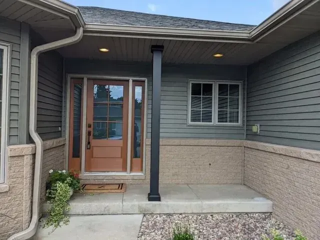 Entryway with brown door, dark pillar, grey siding, stone accents, and small window.