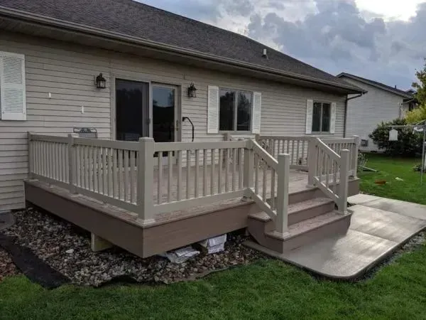 A beige wooden deck with railing and steps next to a house with a sliding door, concrete path, and green lawn.