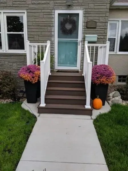 Front entrance with steps and white railings. Brown steps lead to a teal door with a wreath. Purple and orange flowers in black planters. Pumpkin on the ground.