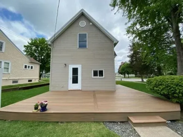 Back view of a tan house with a large wooden deck. A white door and small windows are visible. Green grass and trees surround it.