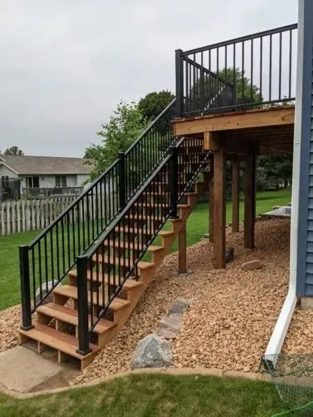Wooden deck with black railing and stairs leading to the ground, surrounded by brown rocks and a green lawn.