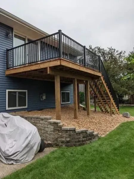Wooden deck with black railing and stairs, attached to a blue house, over a stone retaining wall.