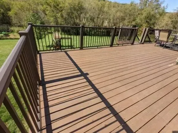 Brown wooden deck with railings, overlooking a grassy yard and trees under a sunny sky.