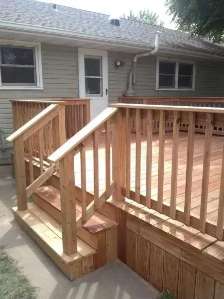 Wooden deck with stairs leading up to it, next to a house with a door and windows.