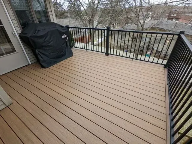 Tan composite deck with black railing and grill cover. Outdoors with a view of trees.