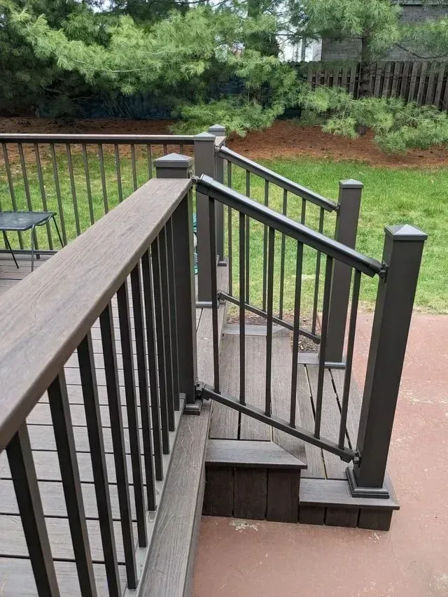 Wooden deck with dark railing and steps, leading to a yard with green grass and trees.
