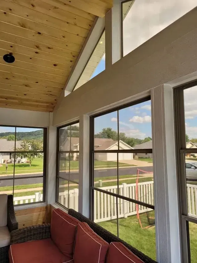Sunroom interior with large windows, wood ceiling, and outdoor view. Red cushions on a wicker sofa.