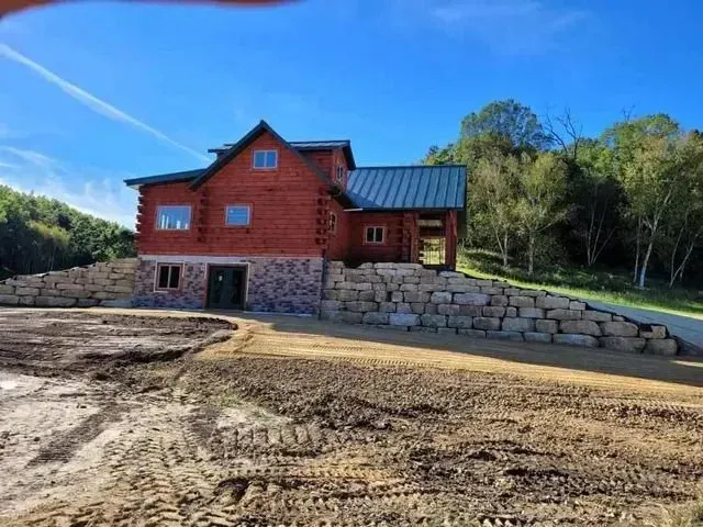 Log cabin with stone foundation and retaining wall, under a clear, blue sky.