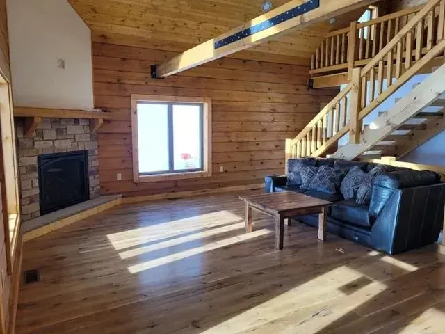 Interior of a log cabin living room with a stone fireplace, wood floors, and a black leather sofa.
