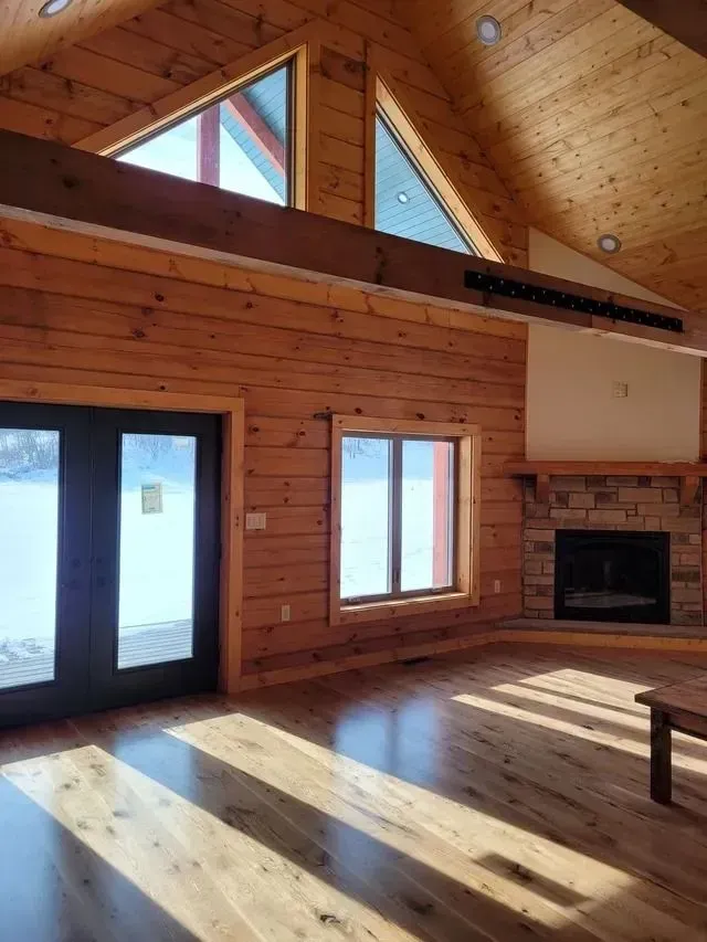 Wooden interior of a home with large windows, fireplace, and hardwood floors. Sunlight streams through.