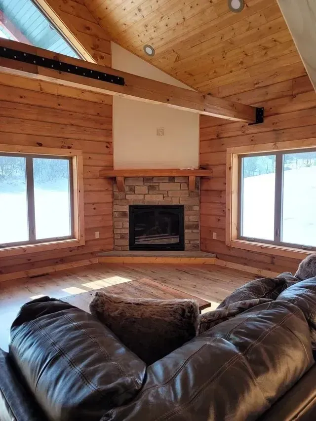 Cozy cabin living room with stone fireplace, wood paneling, large windows, and leather sofa.