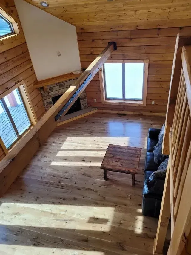 Interior of a log cabin with wooden floors, beams, and large windows. A sofa, table, and fireplace are visible.