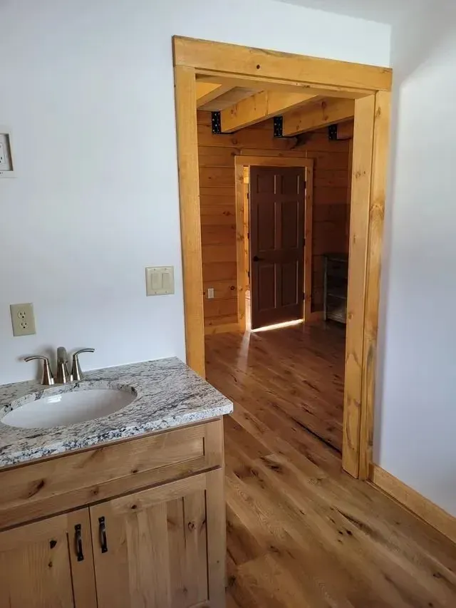 Bathroom with a wooden vanity and a granite countertop, doorway leads to a log cabin interior.