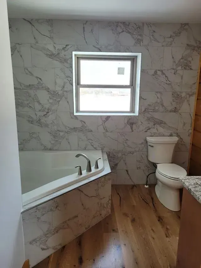 Bathroom with marble-look tile, corner tub, window, and toilet. Wood-look flooring.
