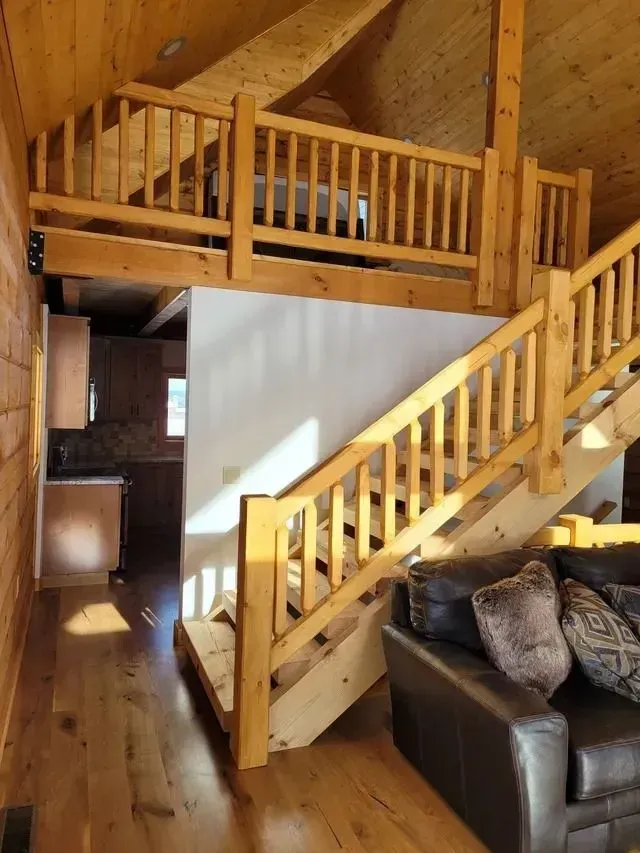 Wooden staircase leading to a loft with a railing, in a cabin-style interior with a brown leather couch.