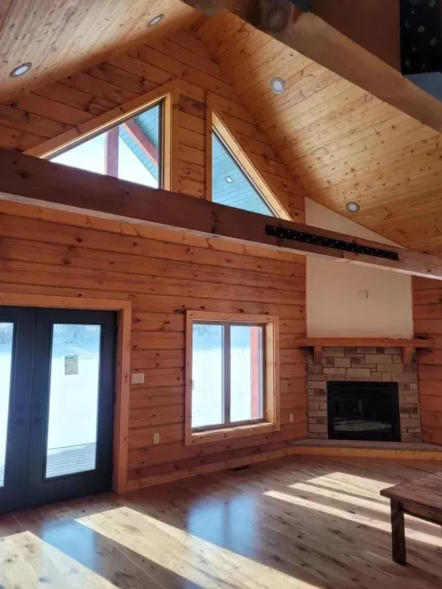 Interior of a wooden cabin with a fireplace, triangular windows, and wooden floors.