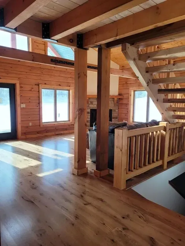 Interior view of a wooden cabin with a staircase, exposed beams, and large windows.