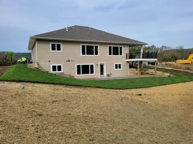 Beige two-story house with black-trimmed windows, a deck, and green lawn on a hillside.
