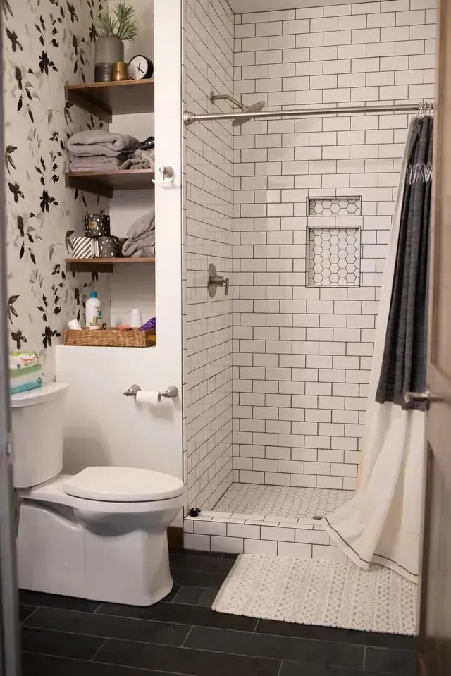 Small bathroom with white subway tile shower, floral wallpaper, open shelving, and black floor tiles.