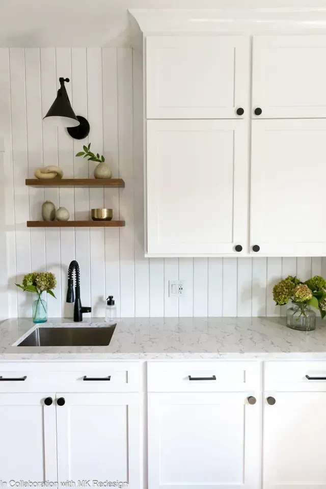 White kitchen cabinets and countertops with black hardware. Black faucet and sconce, wooden shelves.
