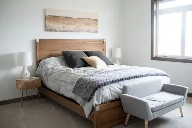 Bedroom with wooden bed, nightstands, bench, window, and abstract art; neutral tones.