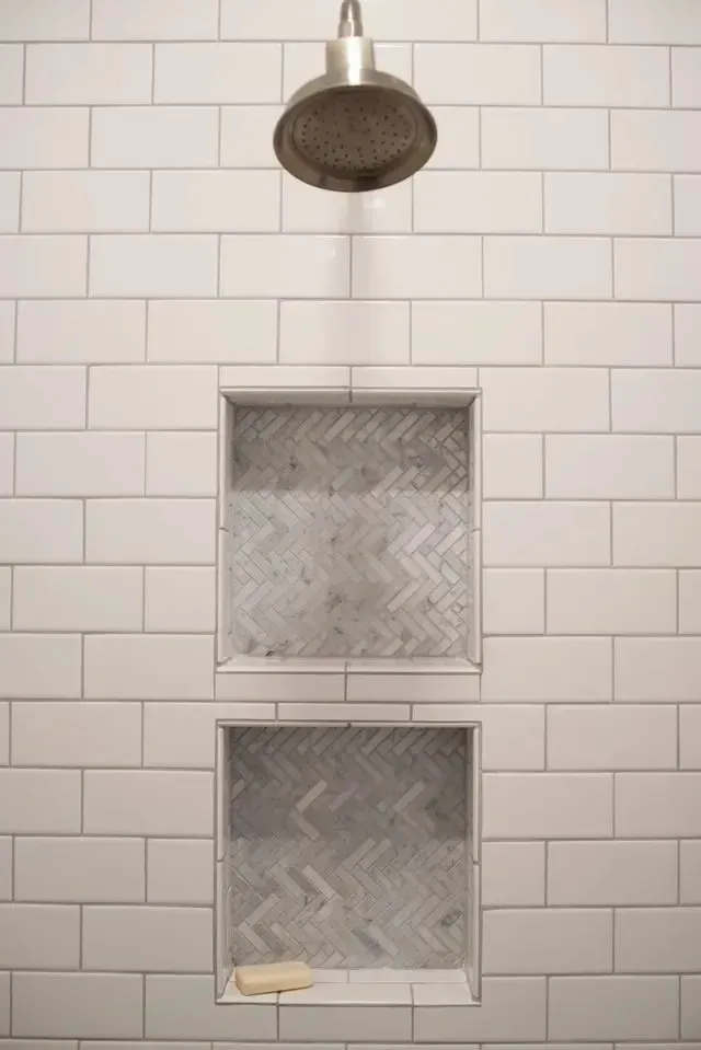 White tiled shower wall with two recessed shelves and a silver showerhead. A bar of soap rests on bottom shelf.