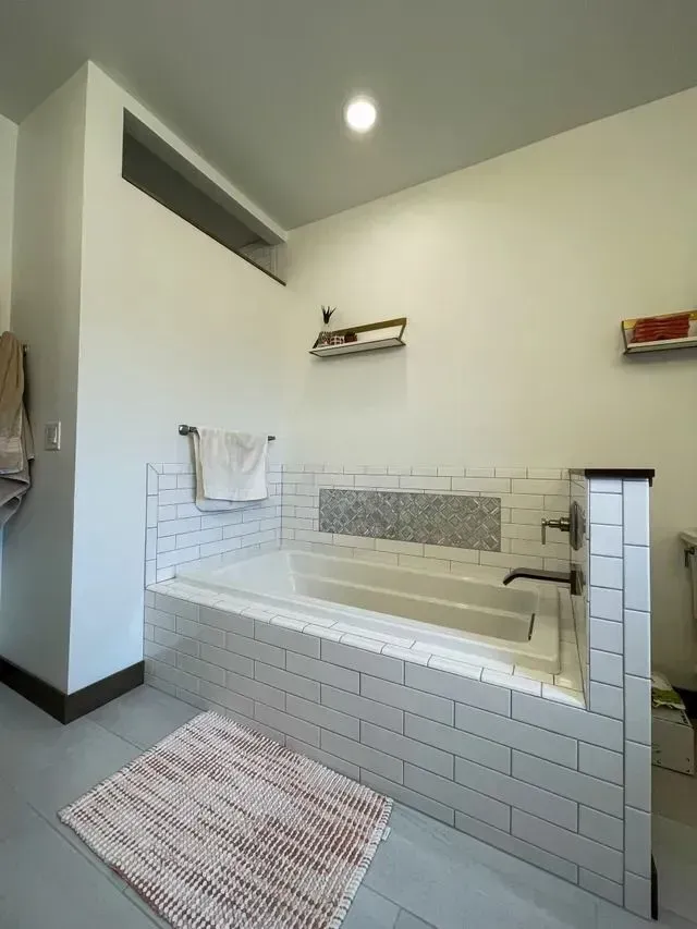 White-tiled bathtub in a bathroom, with a rug, towels, and overhead light.