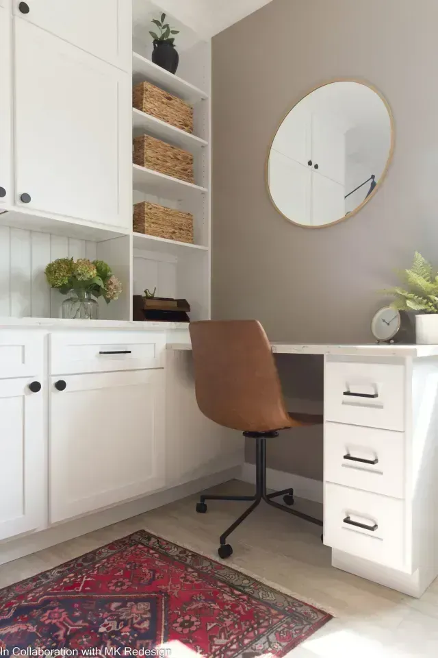 White home office with built-in desk, shelves, and cabinets. Tan chair and oval mirror on the wall.