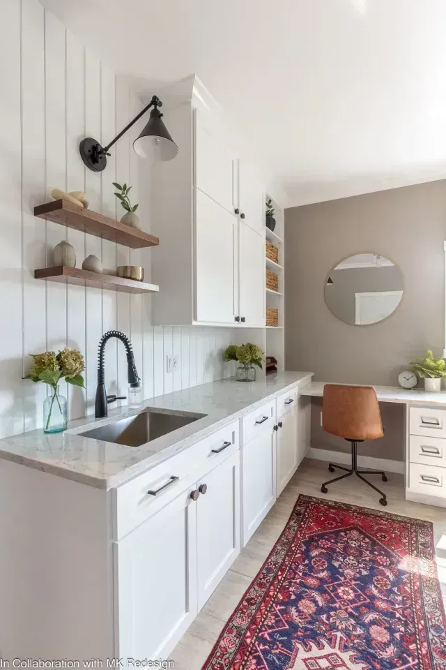 Laundry room with white cabinets, farmhouse sink, wood shelves, and desk area with a rug.