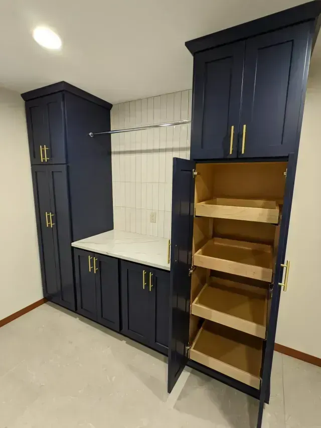 Dark blue pantry cabinets with gold hardware and pull-out shelves, white countertop, and tiled backsplash.