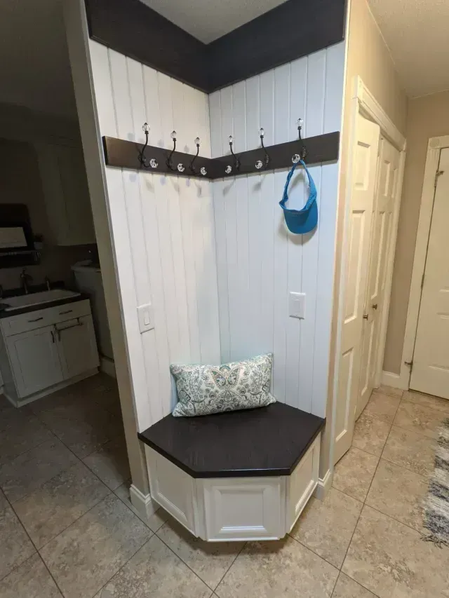Corner entryway with built-in bench, white paneling, dark hooks, and blue hat.