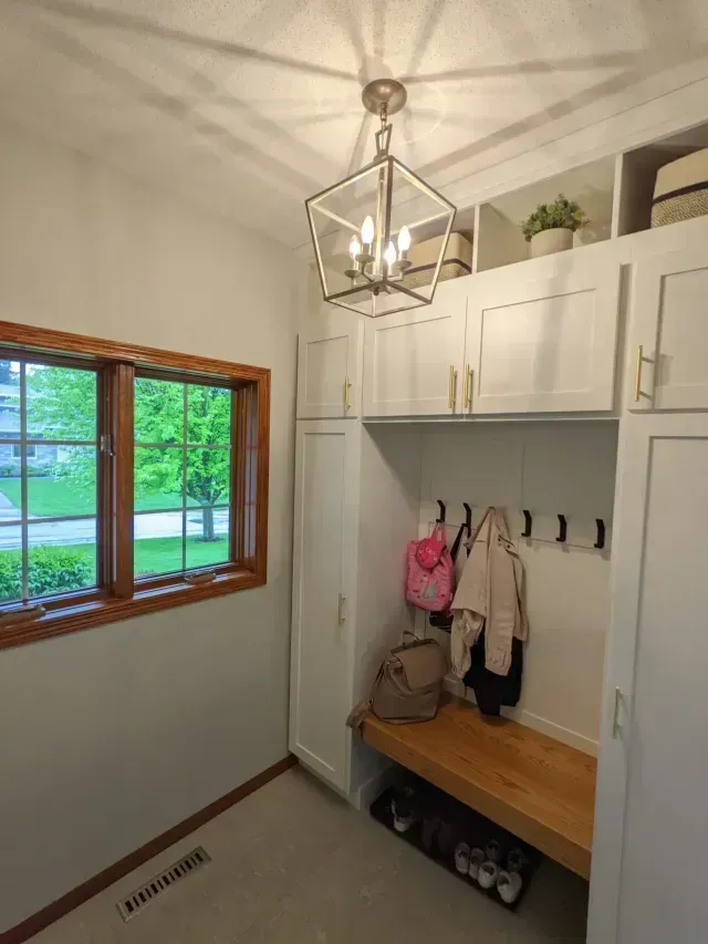 A mudroom with white cabinets, a wood bench, and a window overlooking greenery. A modern light fixture hangs above.