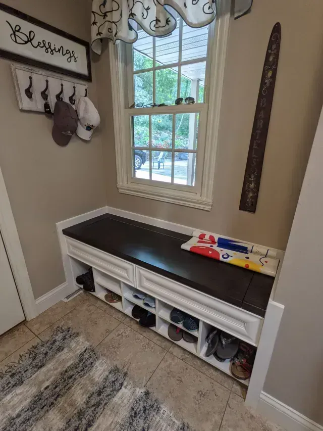 Built-in white bench with shoe storage, dark seat cushion, and a window. Hats hanging on the wall.