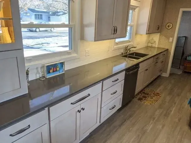 Gray countertop kitchen with light cabinets and dark hardware. Sink under window, dishwasher to the right.