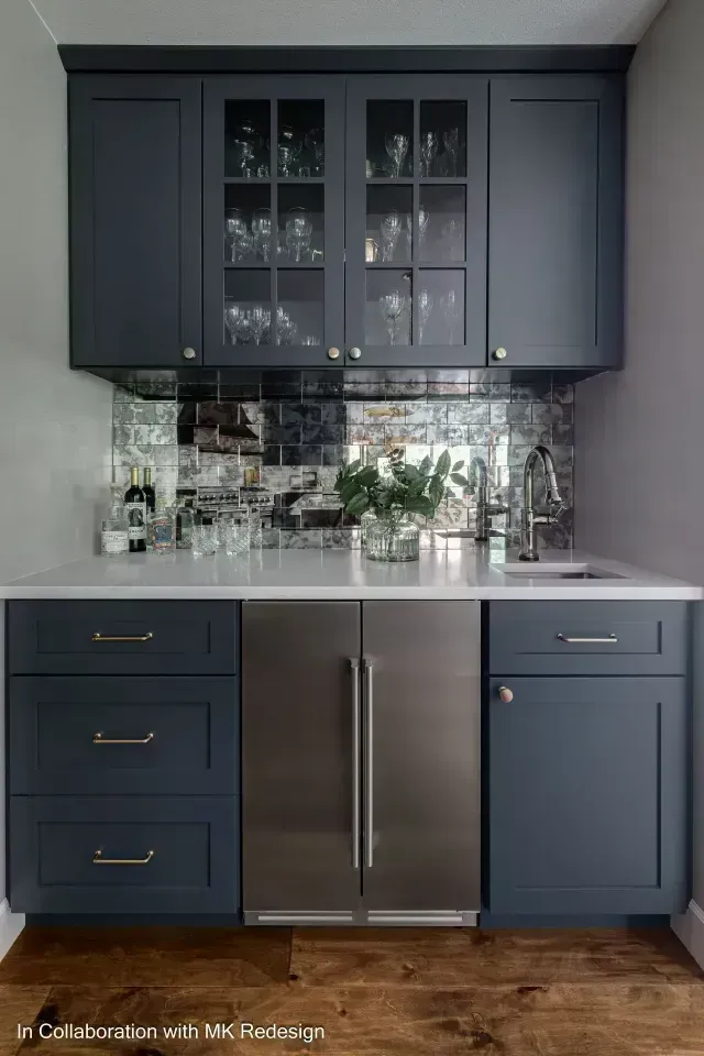 Dark blue home bar with mirrored backsplash, white countertop, and stainless steel refrigerator.