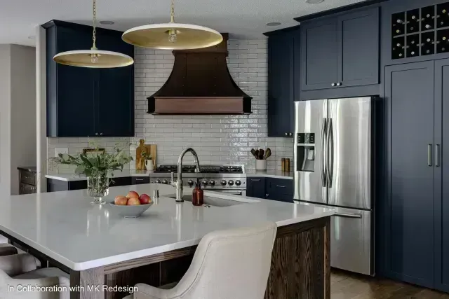 Modern kitchen with navy blue cabinets, stainless steel refrigerator, white countertops, and copper range hood.