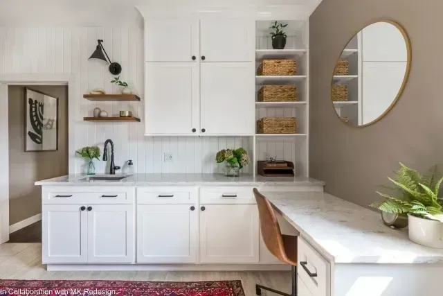 White built-in cabinetry with sink and desk area; light gray walls, open shelving, and a round mirror.