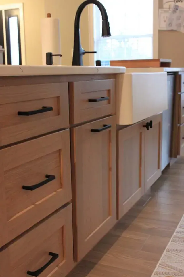 Light wood kitchen cabinets with black hardware, white farmhouse sink, and black faucet.