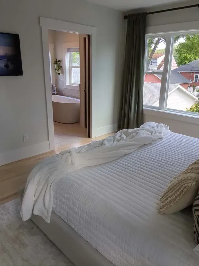 Bedroom with a bed covered in a white quilt, adjacent to a bathroom with a tub, and a window with green drapes.